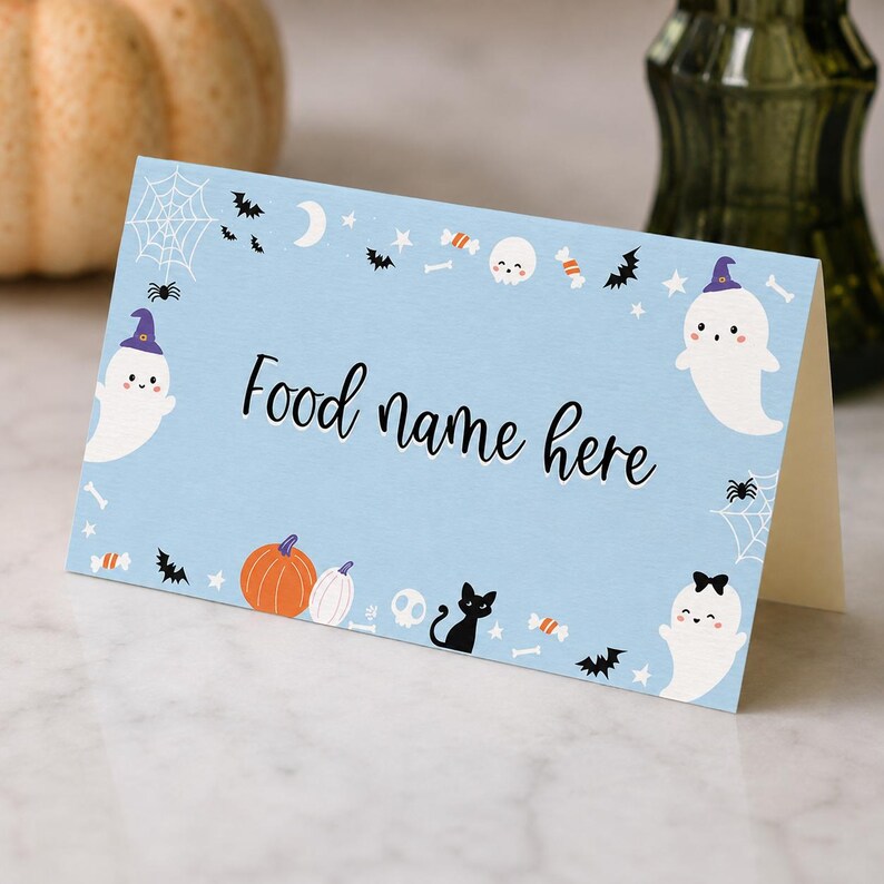 May include: A light blue folded card with the text Food name here in black script. The border features illustrations of white ghosts with hats, orange pumpkins, black cats, bats, spiderwebs, skulls, bones, stars, and candy in a Halloween theme.