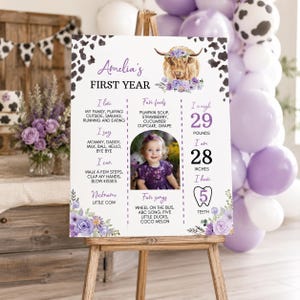 May include: A white milestone birthday sign featuring a highland cow illustration with purple flowers, cow print borders, and text detailing Amelia's first year, including favorite foods, songs, 29 pounds, 28 inches, and 5 teeth.