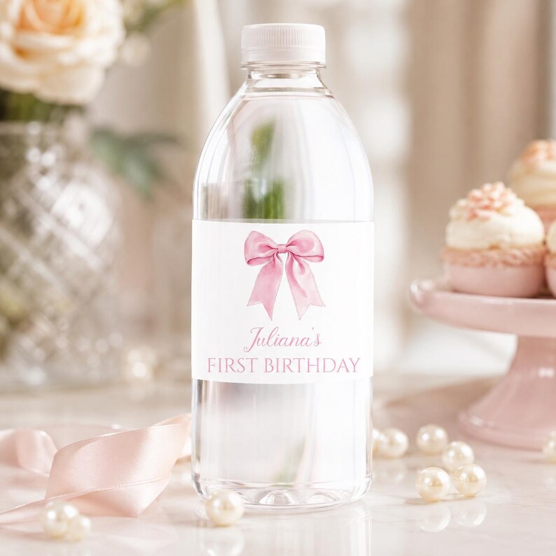 Puede incluir: Una botella de agua de pl&aacute;stico transparente con una etiqueta blanca que presenta una ilustraci&oacute;n de un lazo en acuarela rosa y el texto Juliana's FIRST BIRTHDAY en letras rosas. La botella tiene una tapa de rosca blanca y contiene l&iacute;quido transparente.