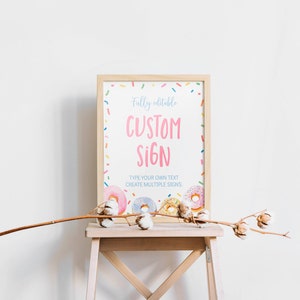 K&ouml;nnte beinhalten: Ein rechteckiges Schild in einem hellen Holzrahmen mit wei&szlig;em Hintergrund, bunten Streuseln, rosa, blauen und gelben Donuts sowie dem Text Fully editable CUSTOM SIGN, TYPE YOUR OWN TEXT, CREATE MULTIPLE SIGNS in Rosa und Blau.