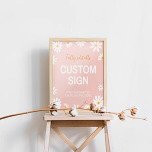 May include: A rectangular sign in a light wood frame featuring a pink background with white daisy illustrations. The text reads: Fully editable CUSTOM SIGN. TYPE YOUR OWN TEXT CREATE MUTIPLE SIGNS. A dried cotton branch rests in front of the frame.
