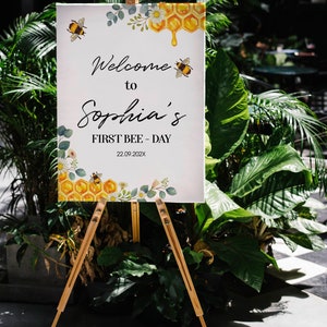 May include: A white sign on a wooden easel, with the text "Welcome to Sophia's FIRST BEE-DAY 22.09.202X". The sign features bee, honeycomb, and floral illustrations. The sign is surrounded by green plants. The easel is made of light brown wood.