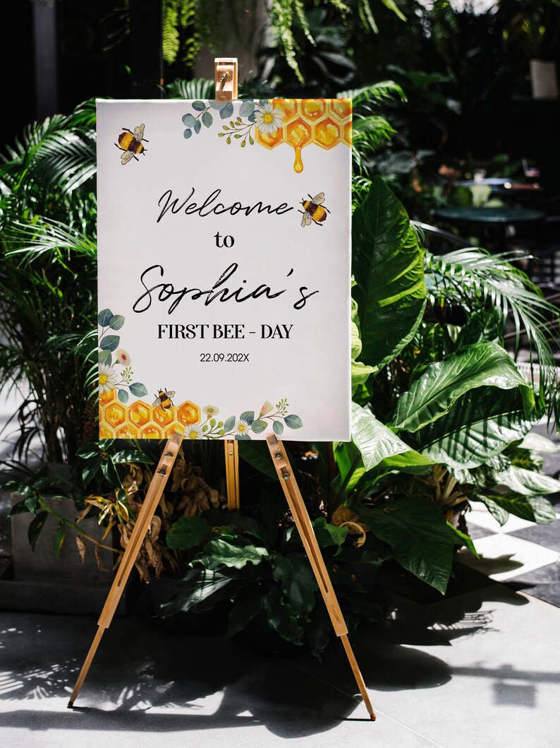 May include: A white sign on a wooden easel, with the text "Welcome to Sophia's FIRST BEE-DAY 22.09.202X". The sign features bee, honeycomb, and floral illustrations. The sign is surrounded by green plants. The easel is made of light brown wood.