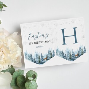 Puede incluir: Un conjunto de dos pancartas de papel con una escena de bosque invernal en acuarela con ciervos, copos de nieve y el texto Easton's 1ST BIRTHDAY 20.12.202X en un lado y una letra H azul grande en el otro, impreso en cartulina blanca.