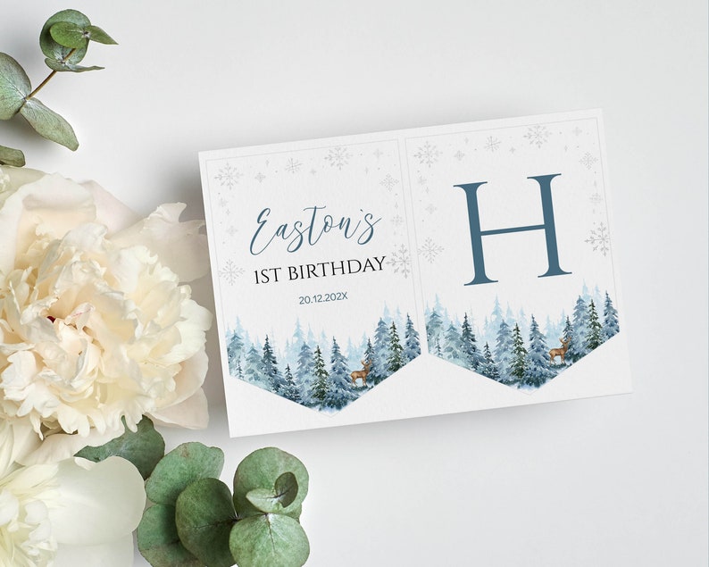 Puede incluir: Un conjunto de dos pancartas de papel con una escena de bosque invernal en acuarela con ciervos, copos de nieve y el texto Easton's 1ST BIRTHDAY 20.12.202X en un lado y una letra H azul grande en el otro, impreso en cartulina blanca.