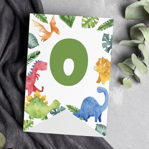 K&ouml;nnte beinhalten: Ein wei&szlig;es, rechteckiges Wimpelf&auml;hnchen mit einem gro&szlig;en gr&uuml;nen Buchstaben O in der Mitte, umgeben von Aquarellillustrationen eines roten Dinosauriers, eines gelben Triceratops, eines blauen Langhalsdinosauriers, eines orangefarbenen Flugsauriers und gr&uuml;nen Bl&auml;ttern.