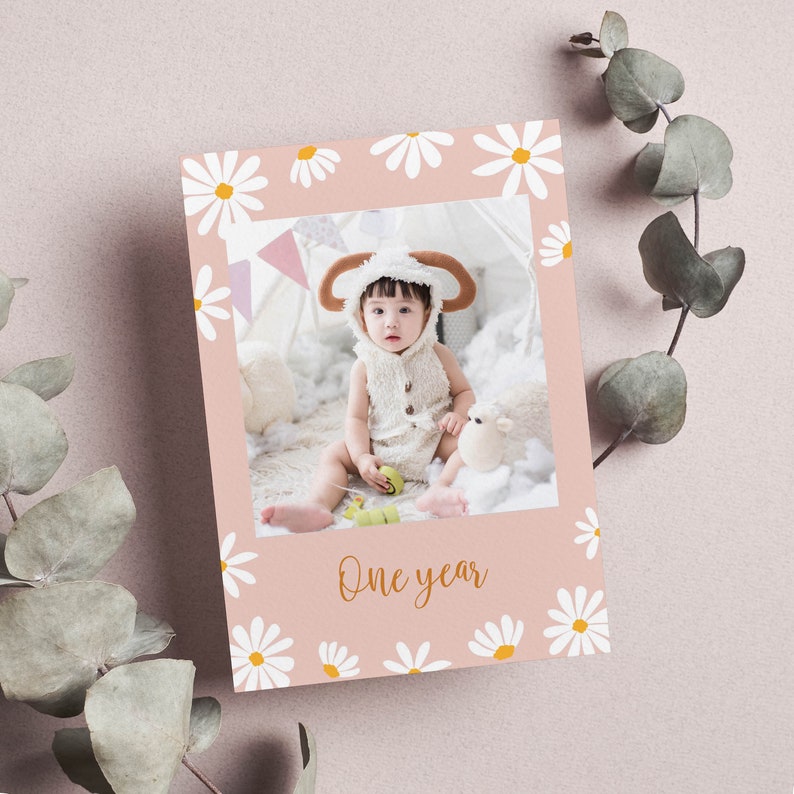 Puede incluir: Una tarjeta rosa con un borde blanco de margaritas. La tarjeta tiene una foto de un beb&eacute; con un disfraz de oveja blanco. El texto "One year" est&aacute; escrito en letras doradas en la tarjeta.
