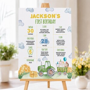 May include: A white birthday milestone sign featuring a green tractor pulling a trailer with animals, yellow text reading JACKSON'S FIRST BIRTHDAY, and sections detailing weight in pounds, height in inches, favorite foods, toys, songs, and words.