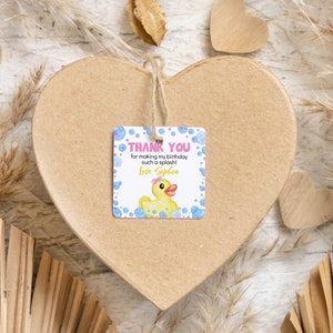 Pu&ograve; includere: Un'etichetta regalo quadrata in carta con una papera di gomma gialla con un fiocco rosa, bolle blu e il testo: THANK YOU for making my birthday such a splash! Love Sophia. L'etichetta &egrave; fissata con uno spago a una grande scatola di cartone a forma di cuore.