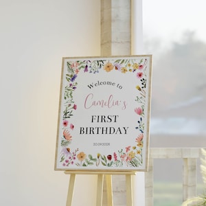 K&ouml;nnte beinhalten: Ein rechteckiges Geburtstagsschild mit einem bunten Aquarell-Blumenrand. Der Text lautet Welcome to Camelia's FIRST BIRTHDAY 20.09.202X. Das Schild steht auf einer hellen Holzstaffelei und pr&auml;sentiert sich vor einem wei&szlig;en Hintergrund.