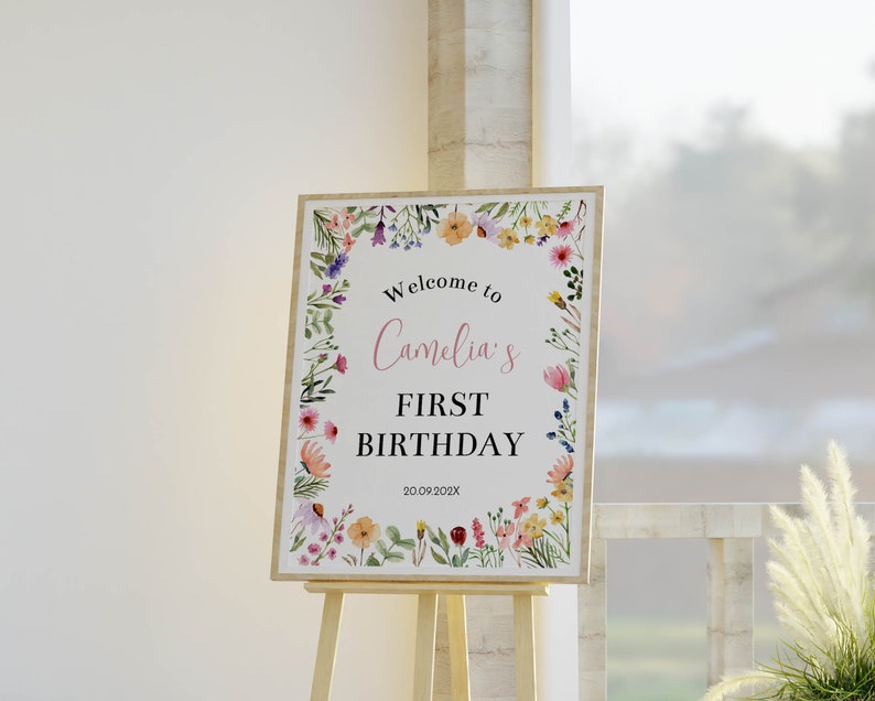 K&ouml;nnte beinhalten: Ein rechteckiges Geburtstagsschild mit einem bunten Aquarell-Blumenrand. Der Text lautet Welcome to Camelia's FIRST BIRTHDAY 20.09.202X. Das Schild steht auf einer hellen Holzstaffelei und pr&auml;sentiert sich vor einem wei&szlig;en Hintergrund.