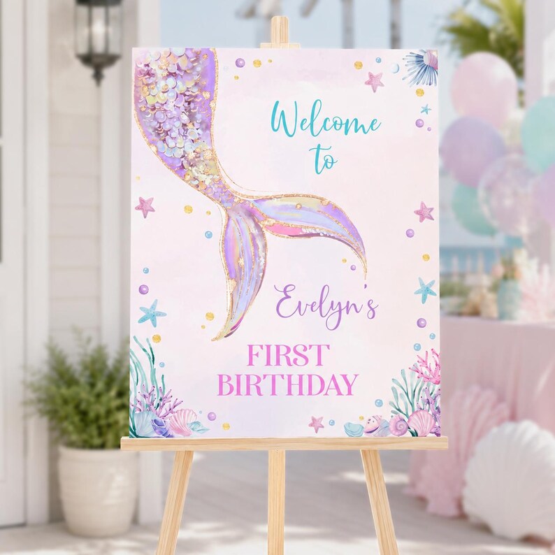 May include: A rectangular sign on a wooden easel featuring a watercolor mermaid tail in shades of purple, pink, and gold. The text reads Welcome to Evelyn's FIRST BIRTHDAY in teal and purple script and block lettering, surrounded by sea life.
