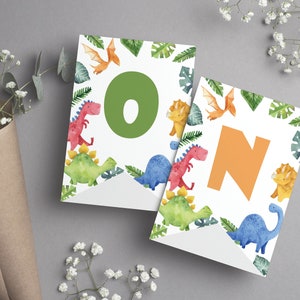 K&ouml;nnte beinhalten: Zwei wei&szlig;e Wimpel mit Aquarellillustrationen verschiedener Dinosaurier und gr&uuml;ner tropischer Bl&auml;tter. Der linke Wimpel zeigt einen gro&szlig;en gr&uuml;nen Buchstaben O, der rechte Wimpel einen gro&szlig;en orangefarbenen Buchstaben N, die zusammen ON ergeben.