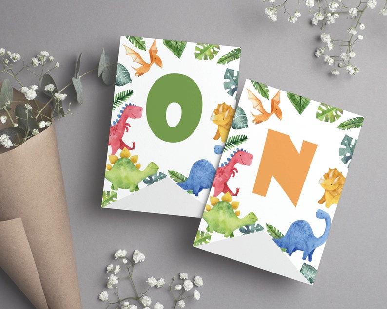 K&ouml;nnte beinhalten: Zwei wei&szlig;e Wimpel mit Aquarellillustrationen verschiedener Dinosaurier und gr&uuml;ner tropischer Bl&auml;tter. Der linke Wimpel zeigt einen gro&szlig;en gr&uuml;nen Buchstaben O, der rechte Wimpel einen gro&szlig;en orangefarbenen Buchstaben N, die zusammen ON ergeben.