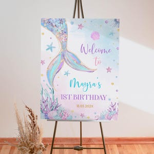 May include: A welcome sign for a first birthday party with a mermaid tail design. The sign is decorated with pastel colors, glitter, and seashells. The text on the sign reads "Welcome to Mayra's 1st Birthday 18.03.2022X".