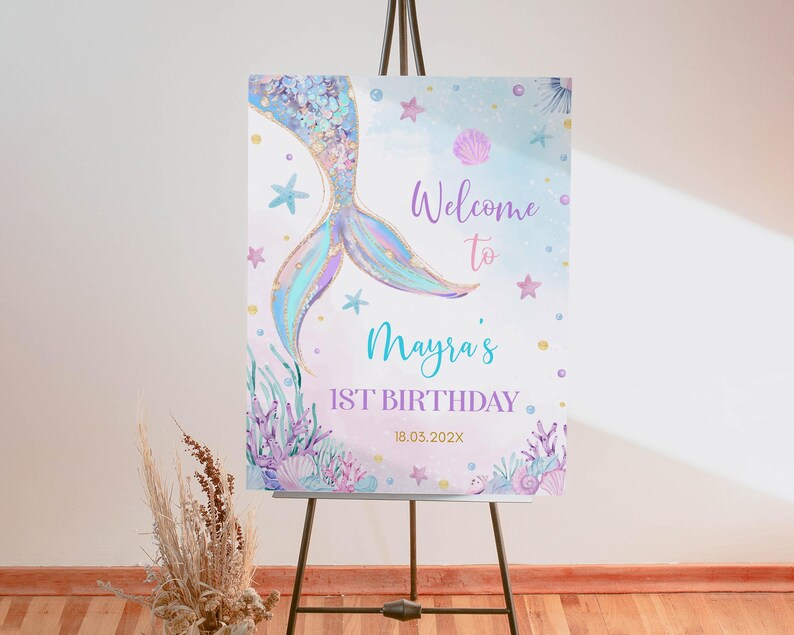 May include: A welcome sign for a first birthday party with a mermaid tail design. The sign is decorated with pastel colors, glitter, and seashells. The text on the sign reads "Welcome to Mayra's 1st Birthday 18.03.2022X".