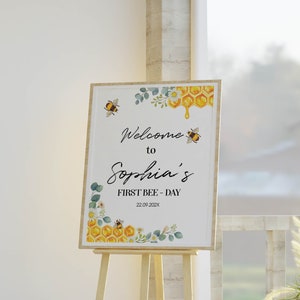 May include: A wooden framed sign on an easel, with the text "Welcome to Sophia's FIRST BEE-DAY" and the date 22.09.202x. The sign has bee, honeycomb, and floral illustrations in yellow, green, and white.