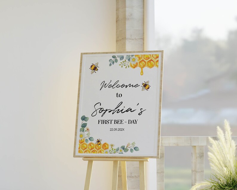 May include: A wooden framed sign on an easel, with the text "Welcome to Sophia's FIRST BEE-DAY" and the date 22.09.202x. The sign has bee, honeycomb, and floral illustrations in yellow, green, and white.