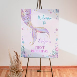 May include: A rectangular sign on a tripod stand featuring a watercolor mermaid tail in purple and gold. The text reads Welcome to Evelyn's FIRST BIRTHDAY. The design includes illustrations of starfish, seashells, and seaweed in pastel colors.