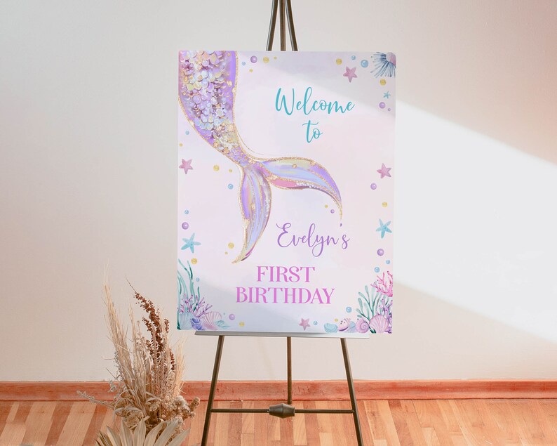 May include: A rectangular sign on a tripod stand featuring a watercolor mermaid tail in purple and gold. The text reads Welcome to Evelyn's FIRST BIRTHDAY. The design includes illustrations of starfish, seashells, and seaweed in pastel colors.