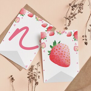 Puede incluir: Dos tarjetas de felicitaci&oacute;n blancas con ilustraciones de fresas y una letra N rosa. Una tarjeta presenta una fresa grande en acuarela, mientras que la otra muestra una letra N rosa grande. Ambas tarjetas est&aacute;n decoradas con fresas peque&ntilde;as.