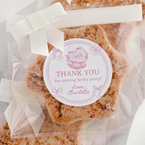 K&ouml;nnte beinhalten: Ein sternf&ouml;rmiger Keks in einer durchsichtigen Plastikt&uuml;te, die mit einem wei&szlig;en Band verschlossen ist. Ein runder Aufkleber auf der T&uuml;te zeigt eine rosa Kuchenillustration, das Wort one und den Text: THANK YOU for coming to my party! Love, Charlotte.