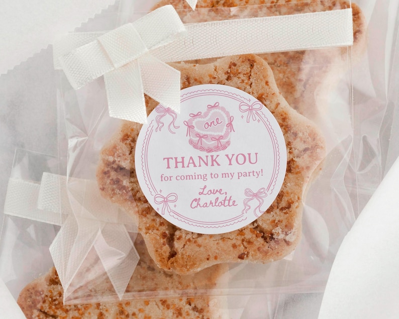 K&ouml;nnte beinhalten: Ein sternf&ouml;rmiger Keks in einer durchsichtigen Plastikt&uuml;te, die mit einem wei&szlig;en Band verschlossen ist. Ein runder Aufkleber auf der T&uuml;te zeigt eine rosa Kuchenillustration, das Wort one und den Text: THANK YOU for coming to my party! Love, Charlotte.