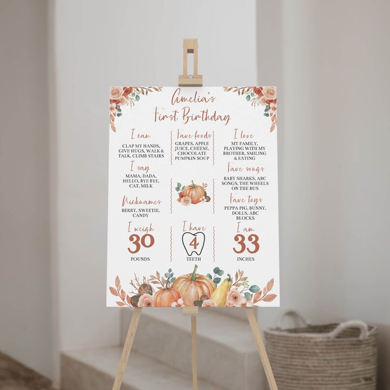 May include: A white birthday milestone sign featuring orange pumpkin illustrations, floral accents, and text detailing Amelia's first birthday facts including weight in pounds, height in inches, favorite foods, songs, toys, and nicknames.