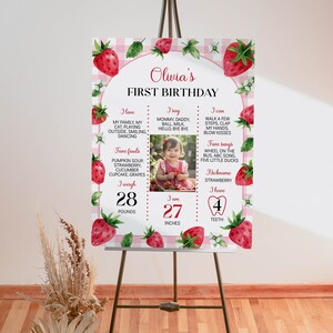 May include: A pink and white first birthday sign with a strawberry theme. The sign includes a photo of a baby girl, the baby's name, Olivia, and details about the baby's first year, including weight, height, favorite foods, and favorite songs.