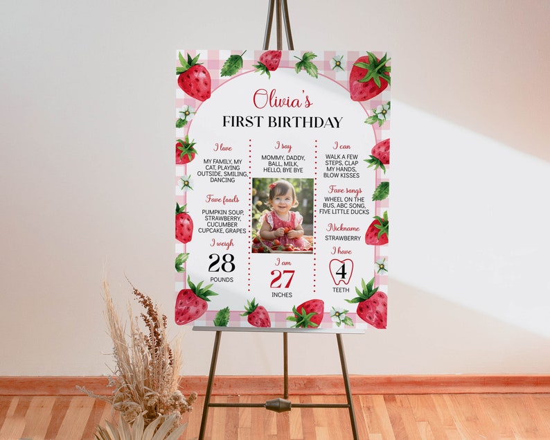May include: A pink and white first birthday sign with a strawberry theme. The sign includes a photo of a baby girl, the baby's name, Olivia, and details about the baby's first year, including weight, height, favorite foods, and favorite songs.