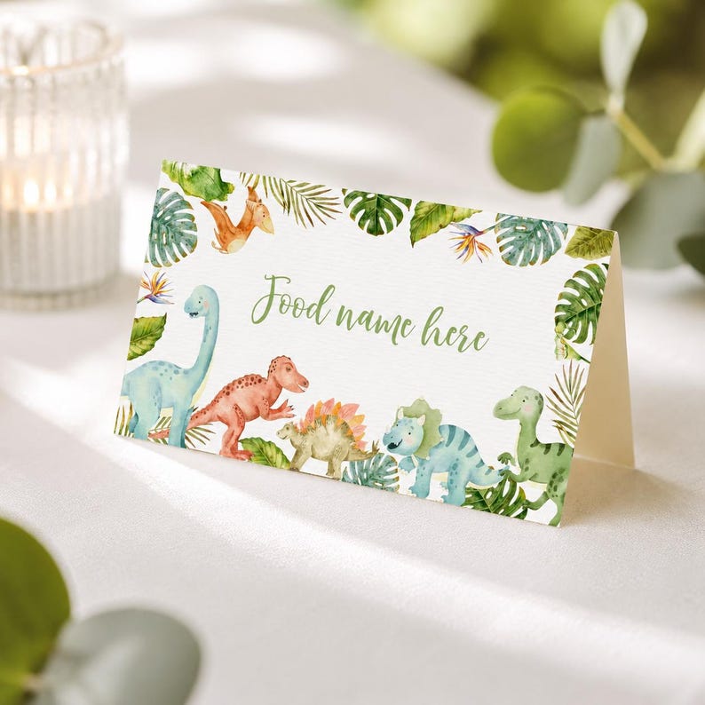 Może przedstawiać: Złożona biała kartka z akwarelową ilustracją r&oacute;żnych dinozaur&oacute;w i tropikalnych zielonych liści. Tekst "Food name here" jest wydrukowany zieloną kursywą na środku kartki, w otoczeniu kolorowych dinozaur&oacute;w.