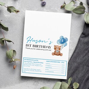 May include: A white rectangular card featuring the text Huson's 1ST BIRTHDAY and Thank you for celebrating with me. It includes a brown teddy bear holding blue balloons with the number 1 and a blue-bordered nutrition facts label.