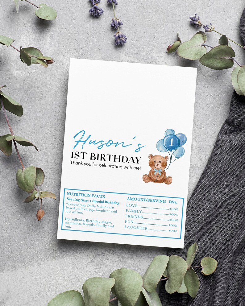 May include: A white rectangular card featuring the text Huson's 1ST BIRTHDAY and Thank you for celebrating with me. It includes a brown teddy bear holding blue balloons with the number 1 and a blue-bordered nutrition facts label.