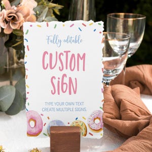K&ouml;nnte beinhalten: Ein rechteckiges wei&szlig;es Schild mit gerissenen Kanten, einem bunten Streuselrand und Donut-Illustrationen am unteren Rand. Der Text lautet Fully editable CUSTOM SIGN TYPE YOUR OWN TEXT CREATE MULTIPLE SIGNS. Das Schild steht in einem braunen Holzblock.