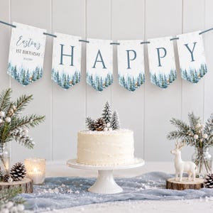 Puede incluir: Un bander&iacute;n con seis gallardetes que deletrean HAPPY y Easton's 1st Birthday 20.12.2024, con un estampado de bosque azul y blanco. Debajo, un pastel glaseado blanco sobre un soporte, rodeado de ramas de pino, pi&ntilde;as y una figura de ciervo.