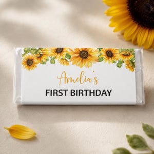 May include: A rectangular candy bar wrapper featuring a white background with a border of yellow sunflowers across the top. The text "Amelia's" is printed in a gold script font, followed by the text "FIRST BIRTHDAY" in bold black capital letters.