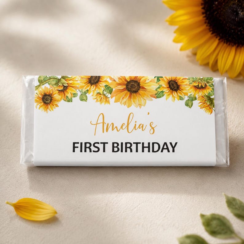 May include: A rectangular candy bar wrapper featuring a white background with a border of yellow sunflowers across the top. The text "Amelia's" is printed in a gold script font, followed by the text "FIRST BIRTHDAY" in bold black capital letters.