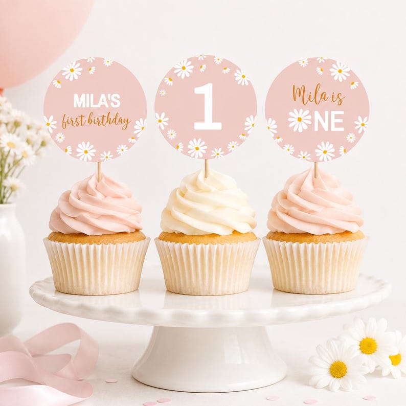 May include: Three cupcakes with light pink frosting on a white stand, each topped with a round paper topper featuring white daisy illustrations. The toppers display the text "Mila's first birthday", the number "1", and "Mila is ONE" in gold and white.
