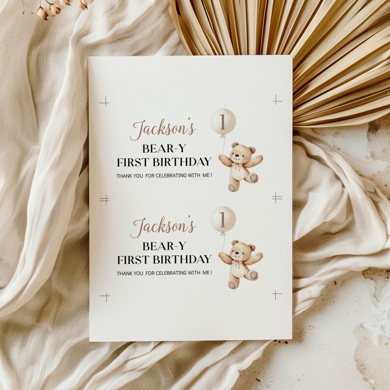 May include: A white rectangular card featuring two identical prints of a brown teddy bear holding a beige balloon with the number 1. The text reads Jackson's BEAR-Y FIRST BIRTHDAY THANK YOU FOR CELEBRATING WITH ME! with black registration marks.