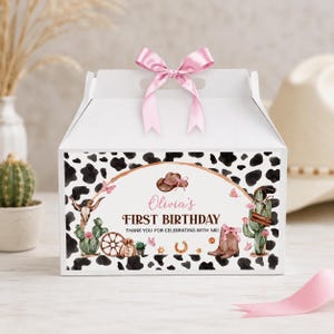 May include: A white gable box with a pink ribbon bow on top, featuring a black and white cow print pattern. The front displays a western-themed graphic with a cowboy hat, cactus, skull, and the text: Olivia's FIRST BIRTHDAY THANK YOU FOR CELEBRATING WITH ME!