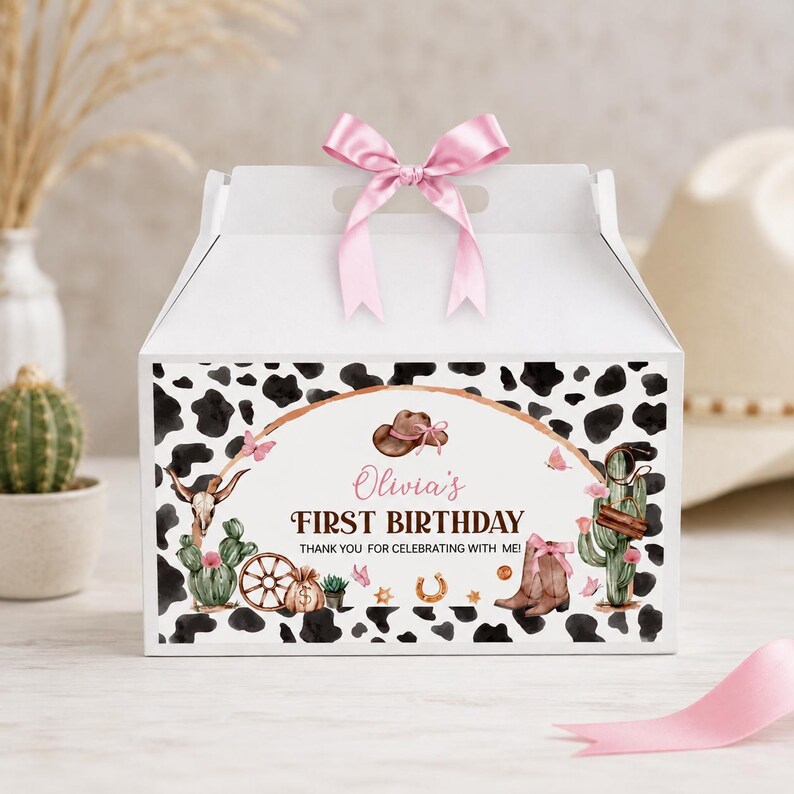 May include: A white gable box with a pink ribbon bow on top, featuring a black and white cow print pattern. The front displays a western-themed graphic with a cowboy hat, cactus, skull, and the text: Olivia's FIRST BIRTHDAY THANK YOU FOR CELEBRATING WITH ME!