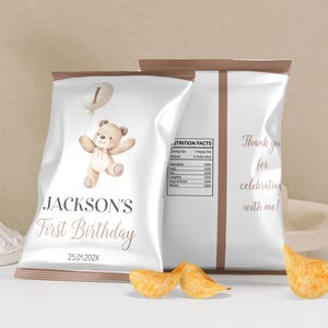 May include: White chip bag with a brown border. The front of the bag has a cartoon illustration of a teddy bear holding a balloon with the number 1 on it. The text "JACKSON'S First Birthday 25.01.202X" is printed on the bag.