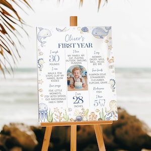 May include: A white milestone board titled Oliver's FIRST YEAR featuring blue watercolor sea creatures like a dolphin, whale, and octopus. It includes text sections for weight, favorite things, and milestones, with a central photo of a child.