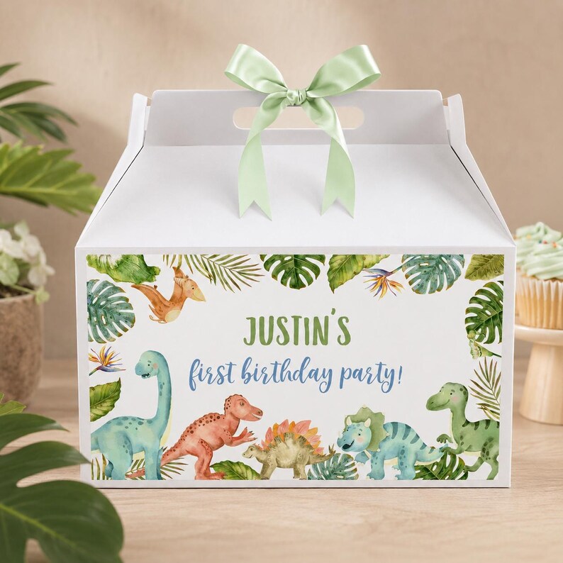 May include: A white cardboard gable box featuring a light green satin ribbon bow on top. The front displays a watercolor illustration of various dinosaurs and tropical leaves with the text Justin's first birthday party in green and blue.