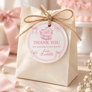 K&ouml;nnte beinhalten: Eine cremefarbene Papiert&uuml;te, die mit braunem Bindfaden und einem runden wei&szlig;en Anh&auml;nger verschlossen ist. Der Anh&auml;nger zeigt rosa Illustrationen von Schleifen und eine herzf&ouml;rmige Torte mit dem Text: one, THANK YOU for coming to my party!, Love, Charlotte.