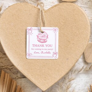 K&ouml;nnte beinhalten: Ein quadratischer, wei&szlig;er Geschenkanh&auml;nger aus Papier mit abgerundeten Ecken, verziert mit rosa Schleifen- und Herzillustrationen, mit dem Text "THANK YOU for coming to my party! Love, Charlotte" und "one" im Herz, befestigt mit einer Schnur an einer herzf&ouml;rmigen Schachtel.