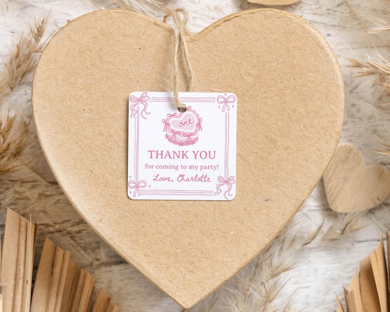 K&ouml;nnte beinhalten: Ein quadratischer, wei&szlig;er Geschenkanh&auml;nger aus Papier mit abgerundeten Ecken, verziert mit rosa Schleifen- und Herzillustrationen, mit dem Text "THANK YOU for coming to my party! Love, Charlotte" und "one" im Herz, befestigt mit einer Schnur an einer herzf&ouml;rmigen Schachtel.