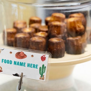 K&ouml;nnte beinhalten: Eine klare Glashaube bedeckt eine Ansammlung kleiner, zylindrischer, dunkelbrauner Backwaren auf einem hellen St&auml;nder. Ein rechteckiges Schild davor zeigt den Text FOOD NAME HERE, verziert mit Illustrationen von Cowboyhut, Stiefeln und Kaktus.
