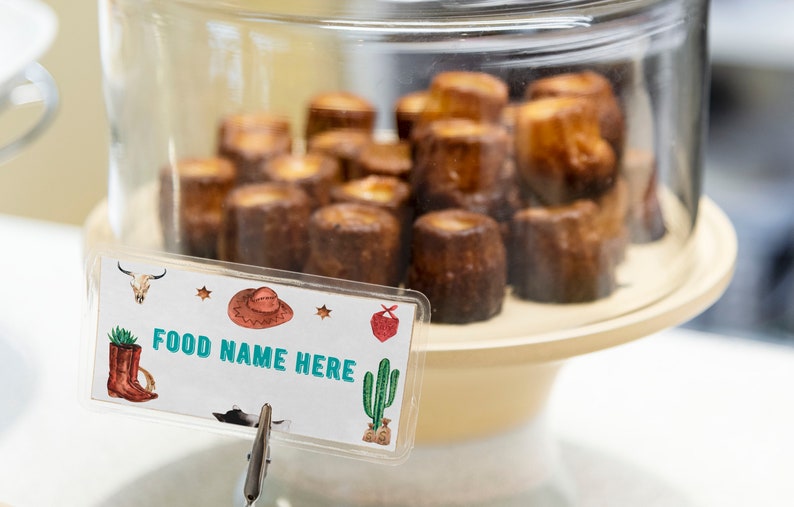 K&ouml;nnte beinhalten: Eine klare Glashaube bedeckt eine Ansammlung kleiner, zylindrischer, dunkelbrauner Backwaren auf einem hellen St&auml;nder. Ein rechteckiges Schild davor zeigt den Text FOOD NAME HERE, verziert mit Illustrationen von Cowboyhut, Stiefeln und Kaktus.