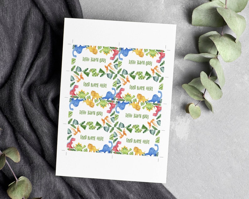 May include: Printable food labels with a dinosaur theme. The labels feature colourful dinosaurs, green leaves, and the text "Food Name Here".